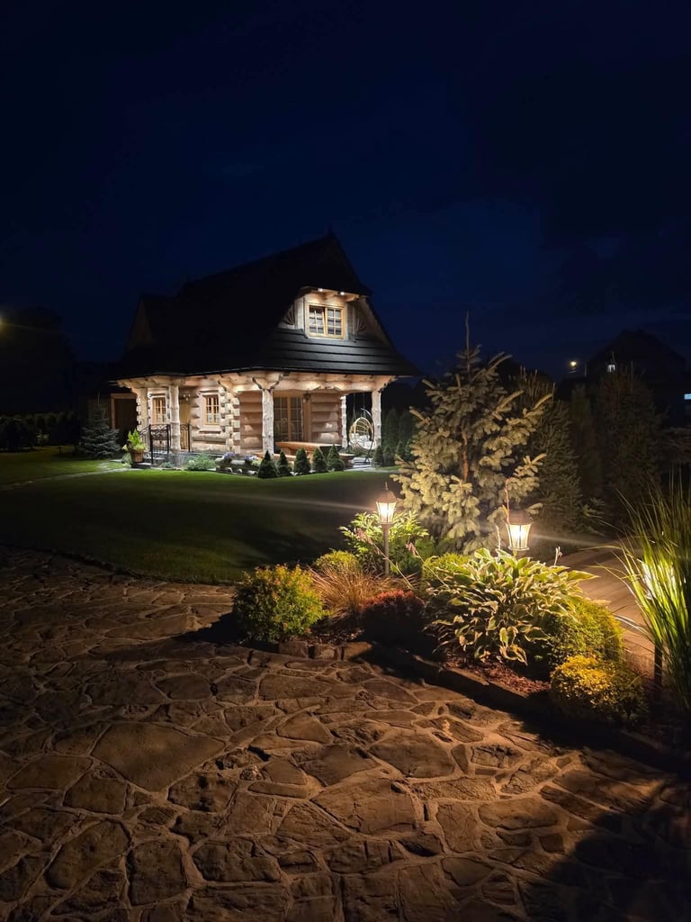 Chata nocą z kamiennym patio, podświetlonym ogrodem i zadbanymi rabatami