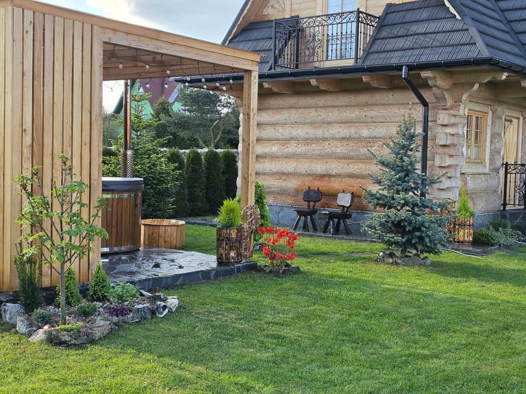 Drewniana pergola przy chatce z zielonym trawnikiem, rabatami kwiatowymi i strefą wypoczynku